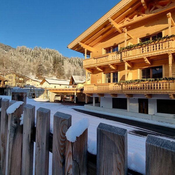 The Farm House exterior in winter, featuring wooden balconies, carports, and snow-covered grounds and fence, with snow-dusted mountains in the background.