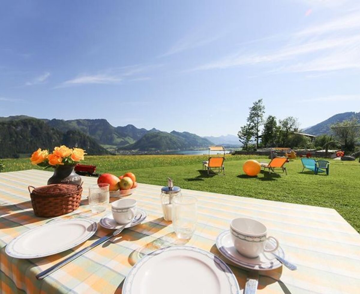 Outdoor breakfast table at the farmhouse with a view of the lake and mountains, surrounded by a lawn with sun loungers.
