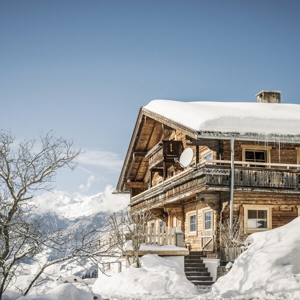 The wooden Farm House exterior, covered in snow, features balconies, icicles from the roof, and snowy mountains in the background.