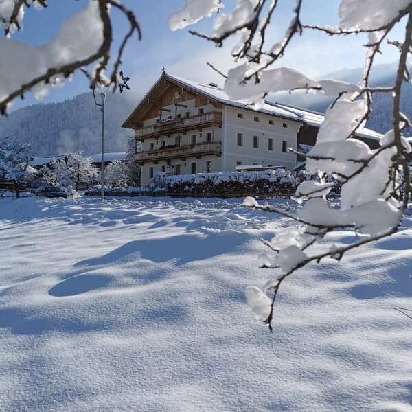 The Farm House exterior in a snow-covered landscape, featuring traditional balconies and framed by snowy branches.