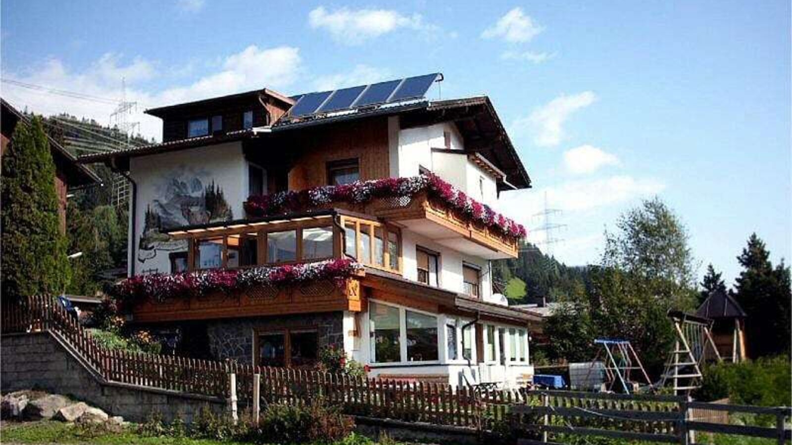 The exterior of the Farm House, featuring balconies with flower boxes, a ground-floor conservatory, solar panels on the roof, and a children's playground in the garden.