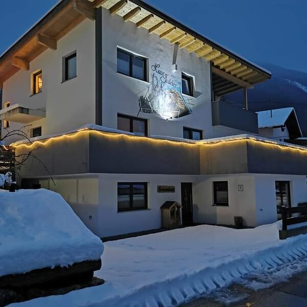 The farm house exterior at night, featuring decorative lighting, snow-covered grounds, and mountains in the background. The farm house exterior at night, featuring decorative lighting, snow-covered grounds, and mountains in the background.