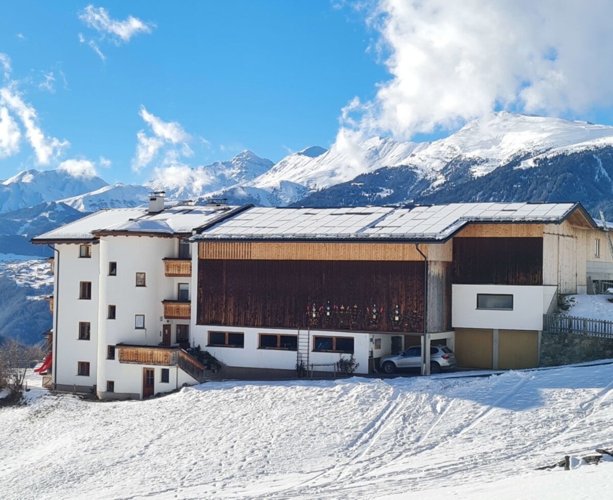 Exterior view of the farmhouse with a white facade, wooden balconies, and a snow-covered roof, surrounded by a winter landscape and mountains.