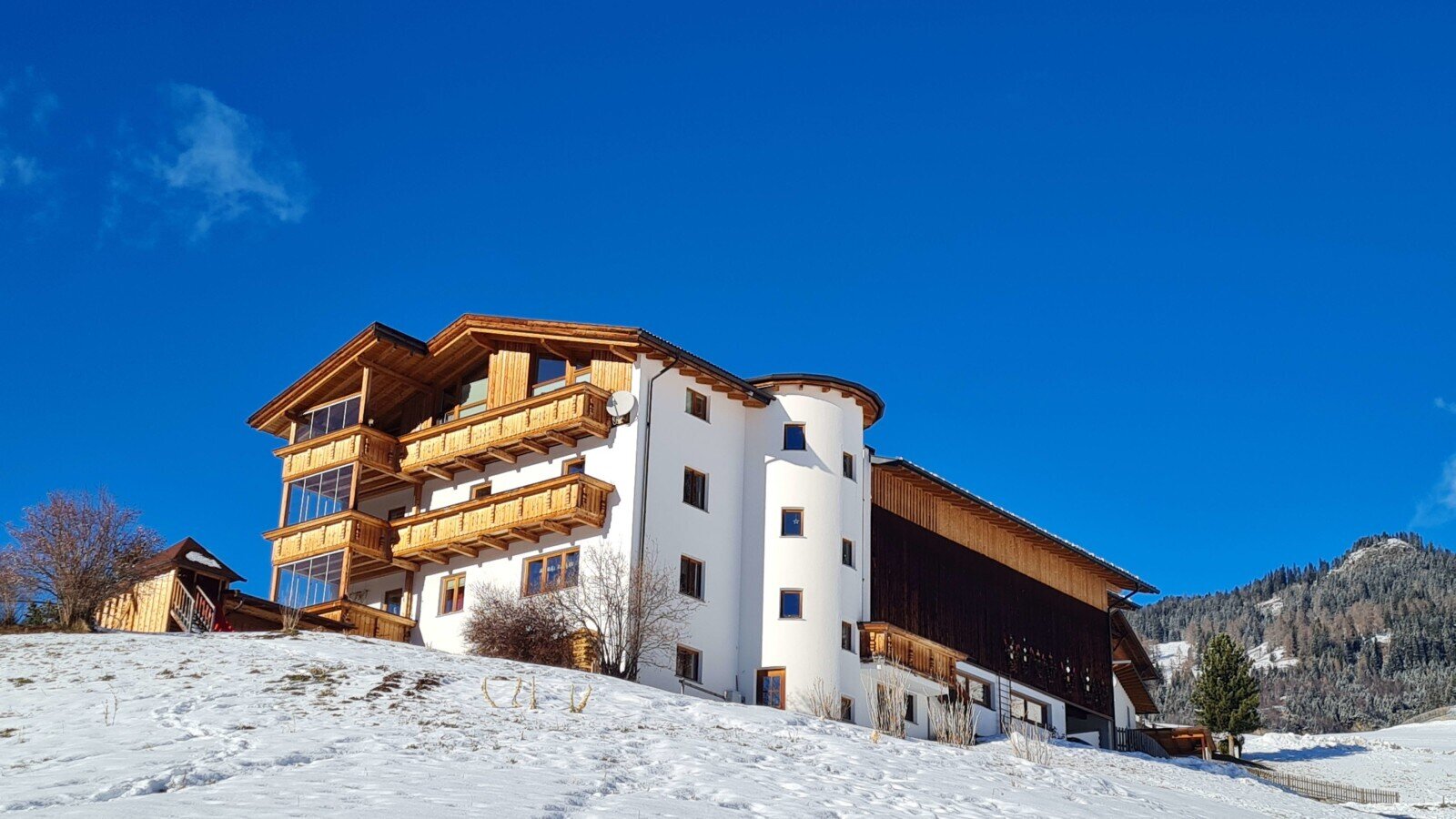The Bauernhof in winter, with a white facade, wooden cladding, and balconies, situated on a snow-covered hill.