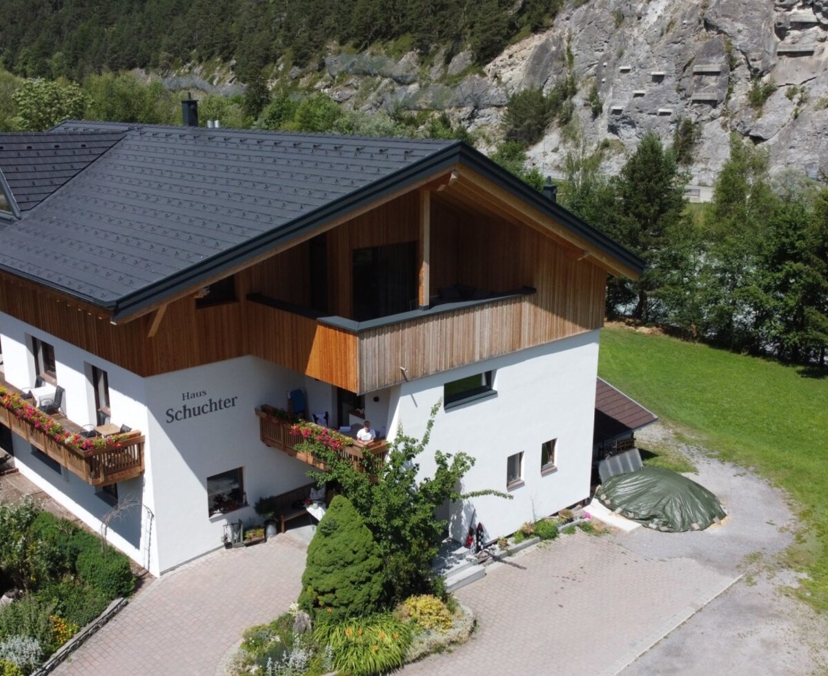 Exterior view of the Bauernhof 'Haus Schuchter' with flower-adorned balconies, surrounded by a garden and mountain scenery.