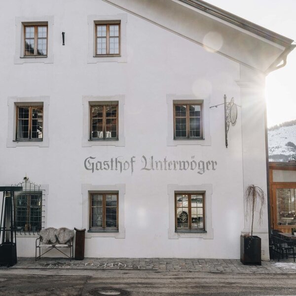 Exterior view of the Bauernhof Gasthof Unterwöger, featuring a white facade, windows, wall paintings, and an outdoor seating area with tables and chairs.