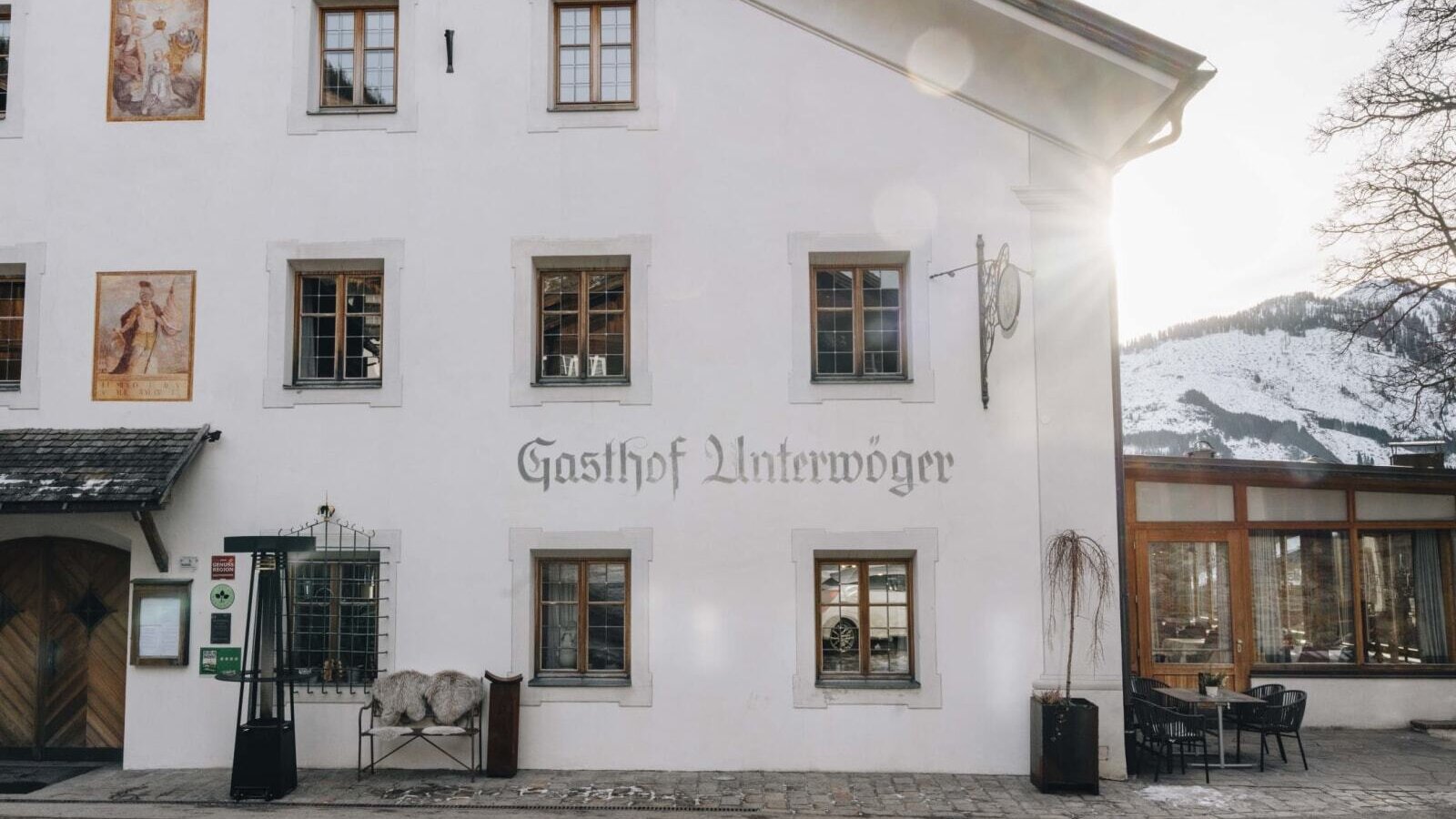 Exterior view of the Bauernhof Gasthof Unterwöger, featuring a white facade, windows, wall paintings, and an outdoor seating area with tables and chairs.