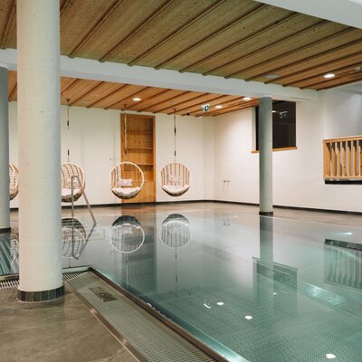 The indoor pool at the farmhouse with several hanging chairs by the poolside and a wooden ceiling.