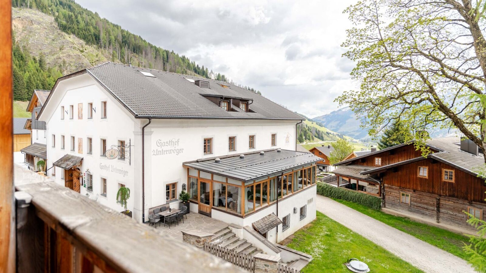 Exterior view of the Bauernhof, featuring a white main building with a glass-enclosed dining area and an outdoor terrace, surrounded by mountains.