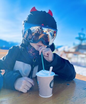 A child in ski goggles and a hat drinks a beverage at an outdoor table, illustrating the family-friendly environment for guests of the Farm House.