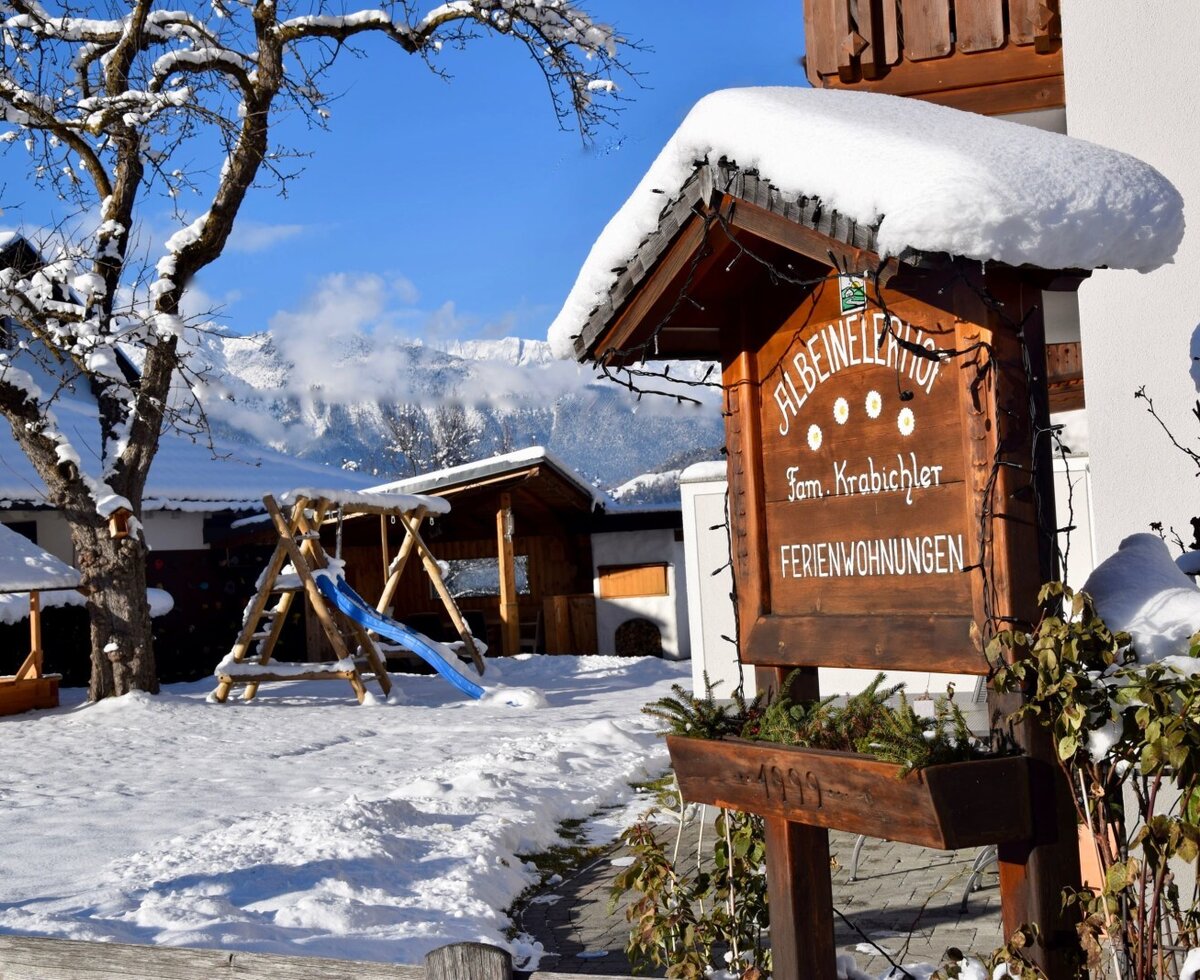 The snow-covered exterior of the Farm House features a wooden sign for the apartments and a children's playground with a slide, set against a mountain backdrop.