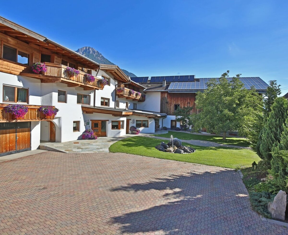 The exterior of the farmhouse, featuring a paved courtyard, a garden with a fountain, wooden balconies with flowers, and mountain views.