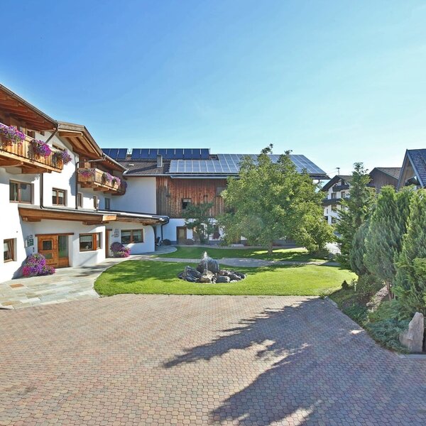 Exterior view of the farmhouse with flower-decorated wooden balconies, a paved courtyard, and a fountain in the garden.