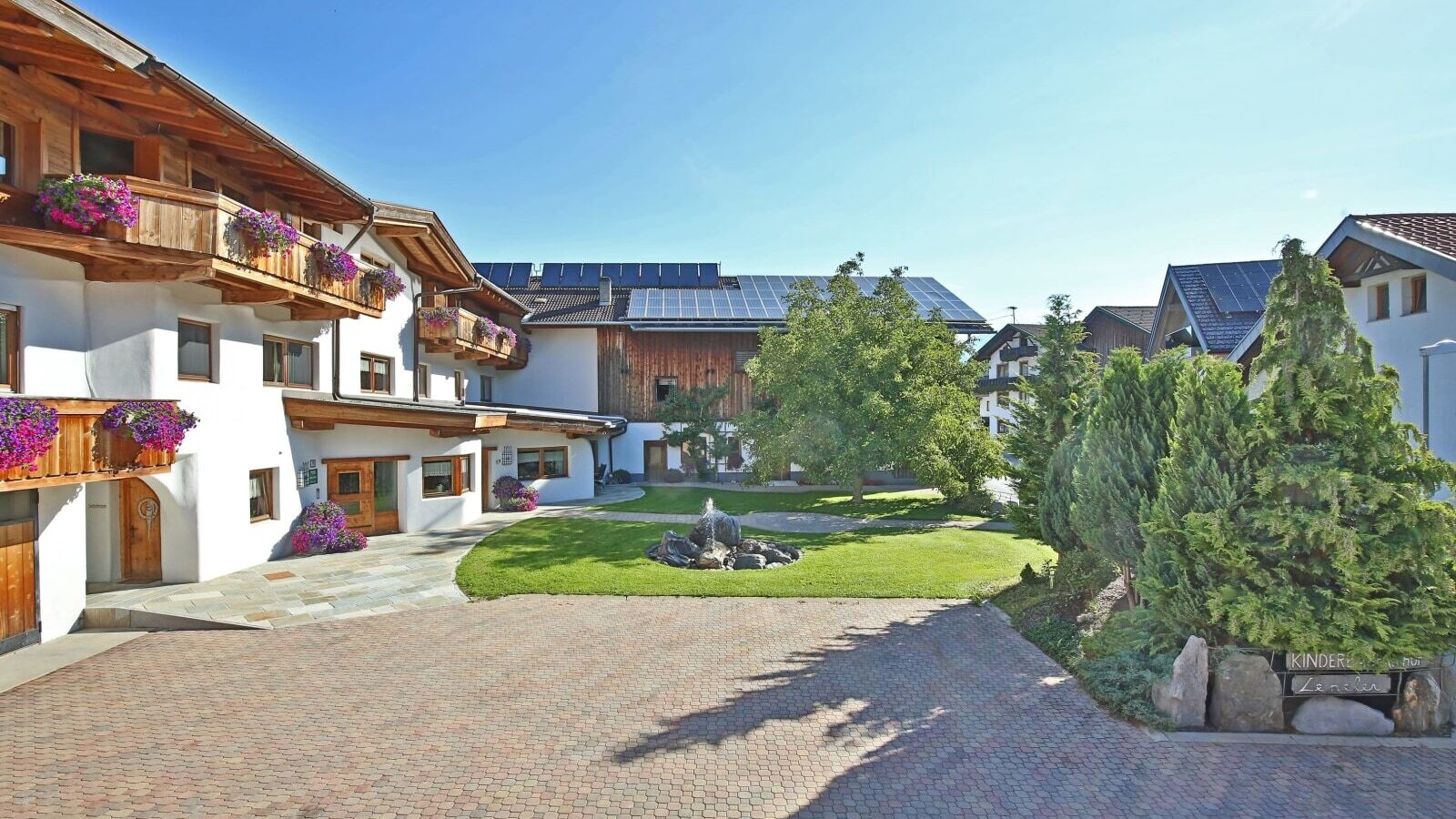 Exterior view of the farmhouse with flower-decorated wooden balconies, a paved courtyard, and a fountain in the garden.