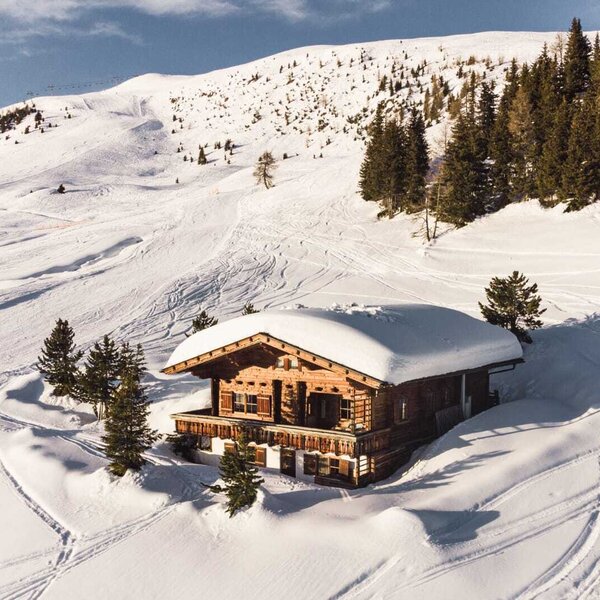 The wooden Alpine Hut features a snow-covered roof and a balcony, situated directly on a ski slope with visible ski tracks.