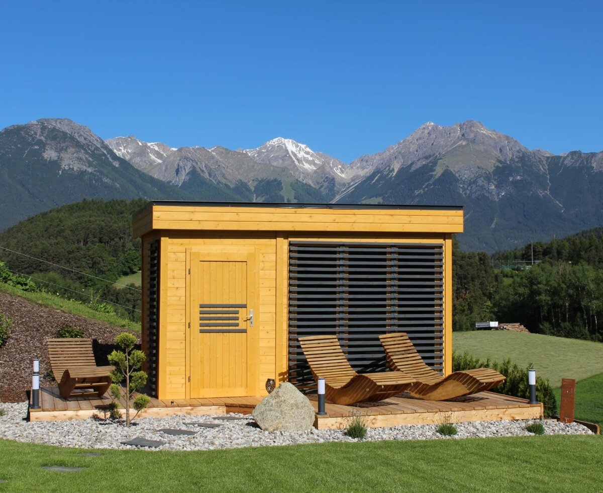 Outdoor sauna of the Ap­par­te­ments with a wooden terrace, lounge chairs, and mountain views.