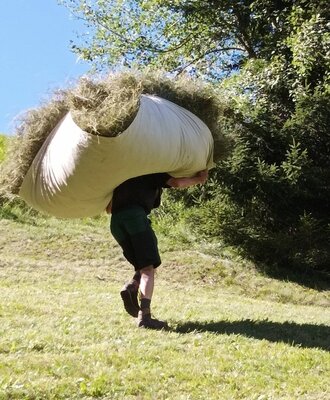 A person carries a large sack of hay across the grassy grounds of the farmhouse.