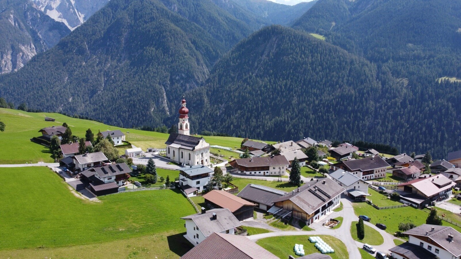 The village surroundings of the Farm House, featuring a central church, traditional buildings, green fields, and forested mountains.