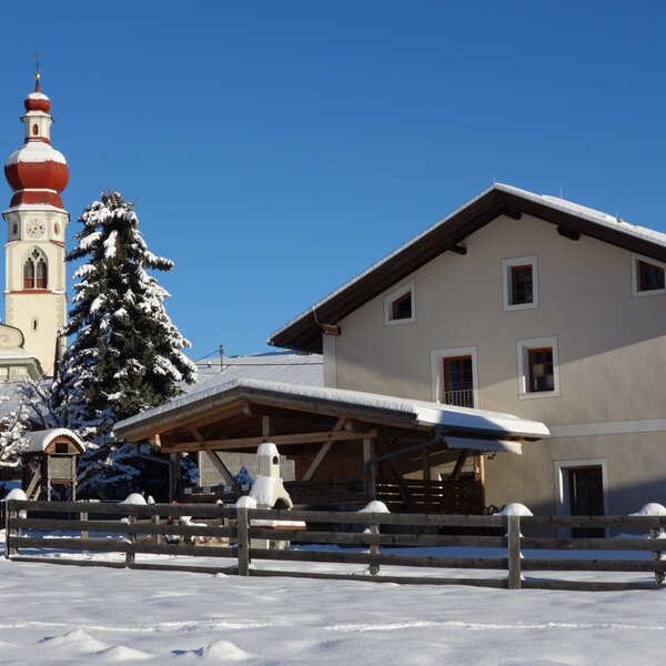 The farm house exterior, featuring a covered outdoor area, is situated in a snowy winter landscape with a church steeple visible in the background.