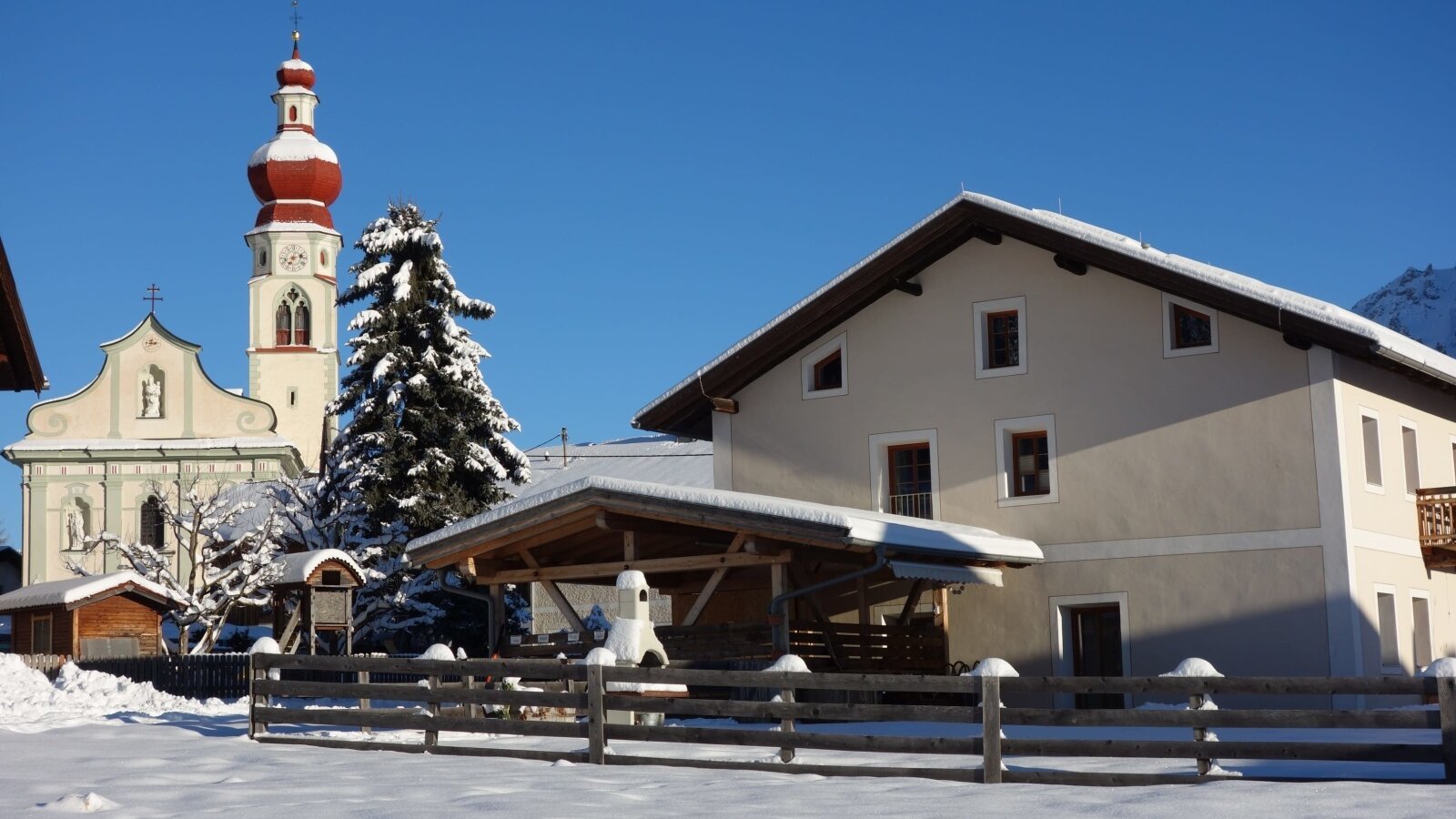 The farm house exterior, featuring a covered outdoor area, is situated in a snowy winter landscape with a church steeple visible in the background.