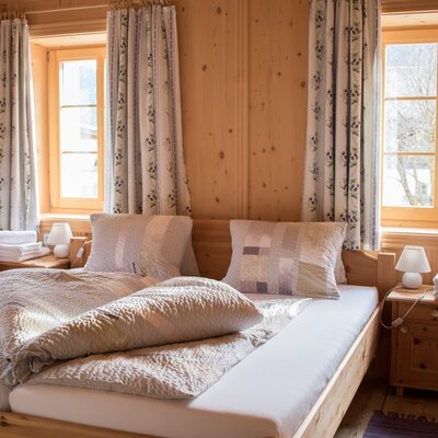 Double bedroom in the farm house with wooden walls, two windows with floral curtains, and bedside tables with lamps.