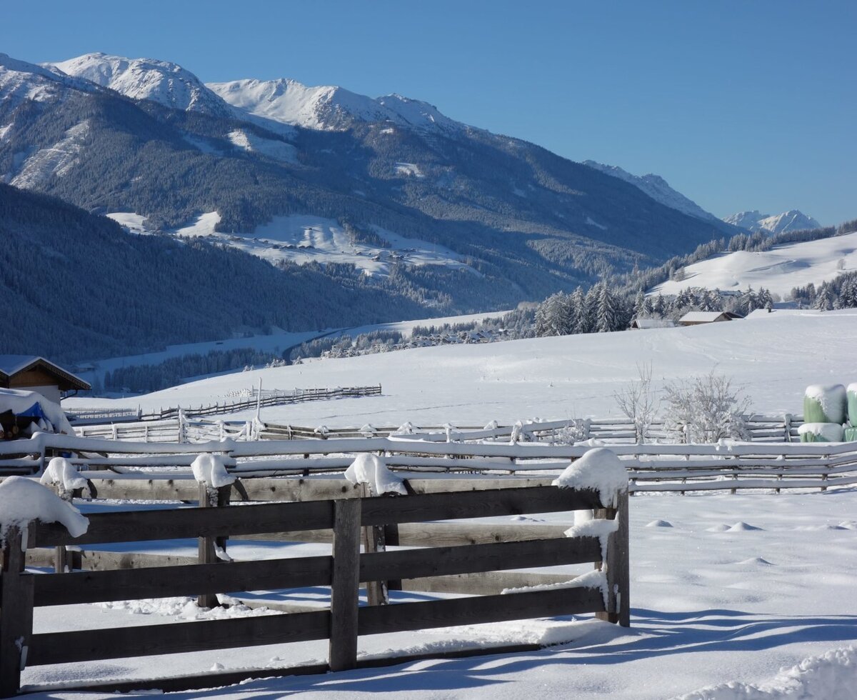 View from the Farm House of snow-covered fields, wooden fences, hay bales, and distant mountains.