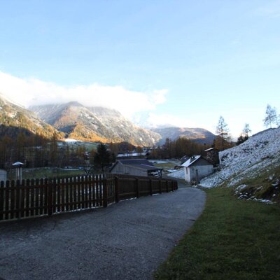 The Farm House is situated in a valley with views of snow-dusted mountains, autumn trees, and a paved path bordered by a wooden fence.