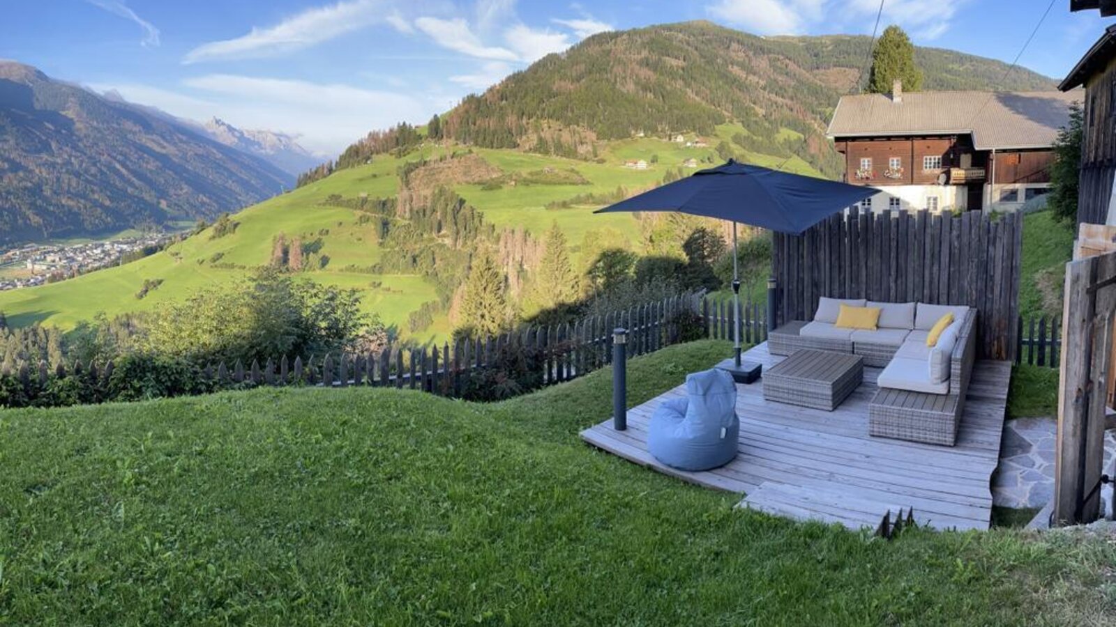 The farm house offers an outdoor wooden deck with sofa seating and an umbrella, providing views of the surrounding mountains and valley.