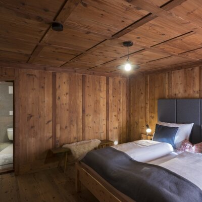 Double bedroom in the farmhouse featuring wooden walls and ceiling, a double bed with a grey headboard, and an en-suite glass-enclosed bathroom.