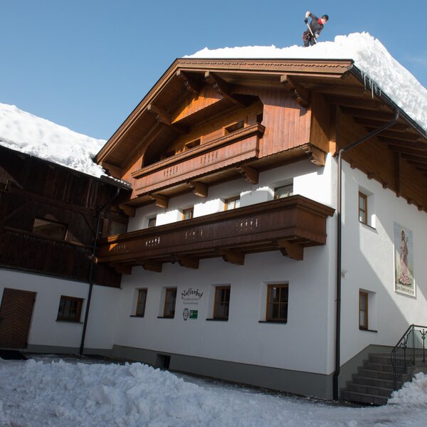 The Farm House exterior in winter, featuring white walls, traditional wooden balconies, and snow on the roof and surrounding grounds.