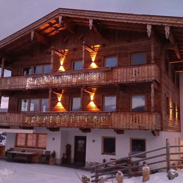 The farmhouse features illuminated wooden balconies and exterior details, set against a snowy landscape with distant mountains.
