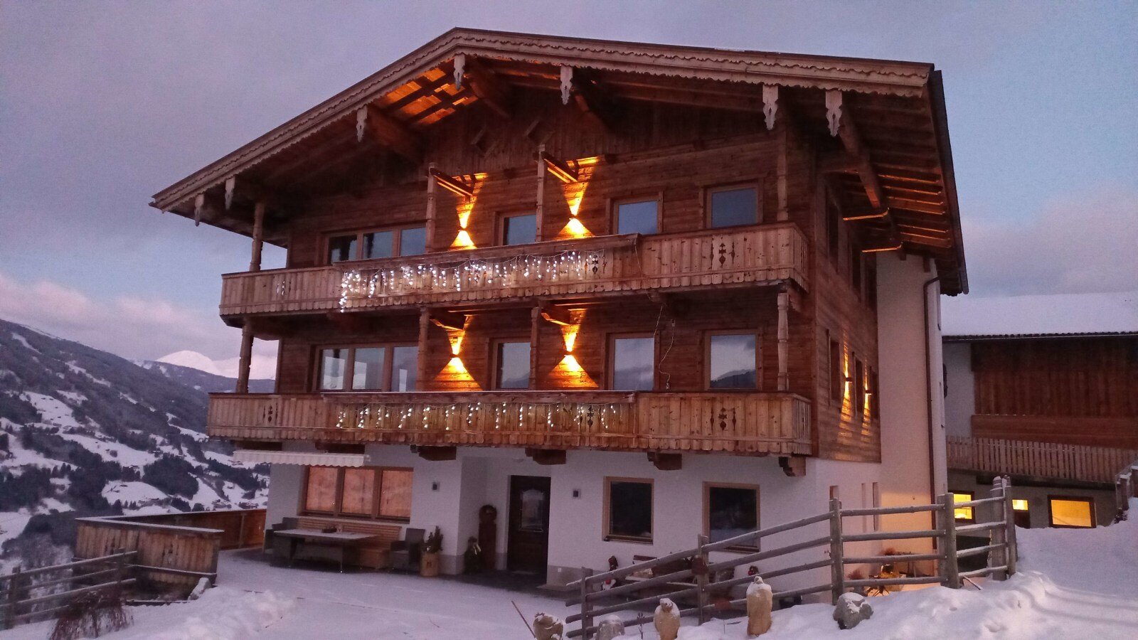 The farmhouse features illuminated wooden balconies and exterior details, set against a snowy landscape with distant mountains.