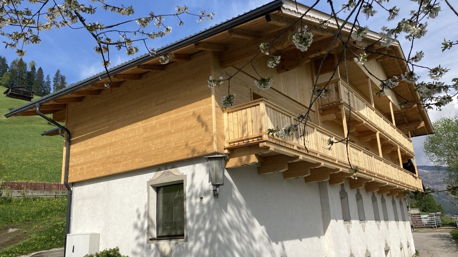 The exterior of the Farm House, featuring wooden balconies and a new wooden upper facade, with blossoming tree branches in the foreground.