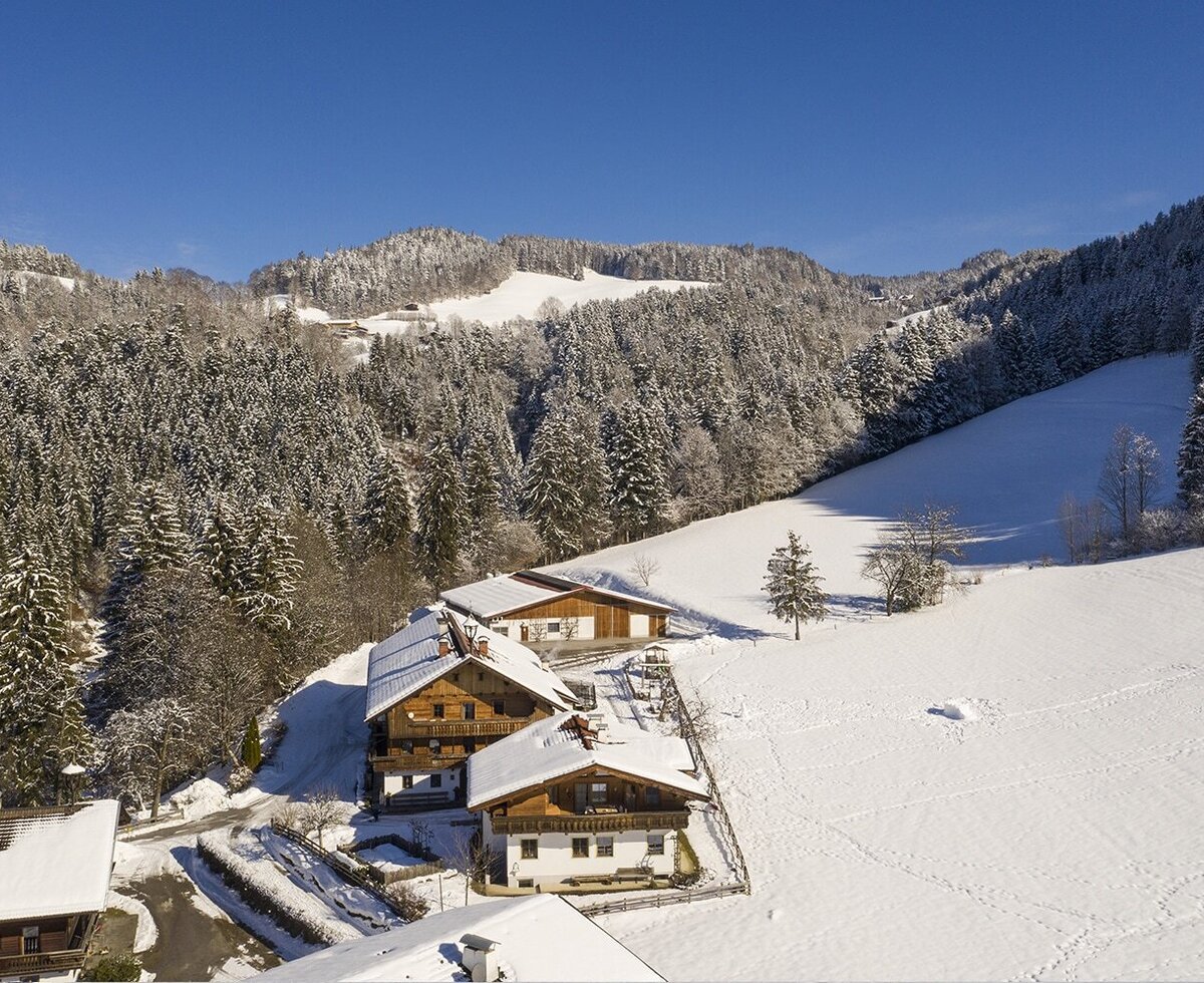 The Farm House property buildings are situated in a snow-covered mountain landscape, surrounded by evergreen forests.