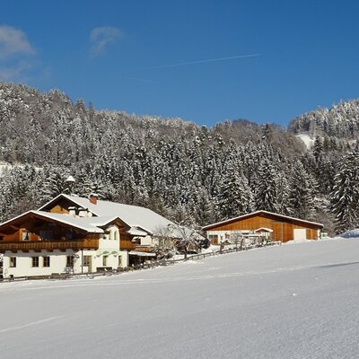 The exterior of the Farm House, featuring traditional architecture and balconies, is situated in a snow-covered landscape with an adjacent outbuilding and surrounding forests.