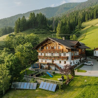 The farmhouse features balconies and a children's playground, set amidst green hills and forests.