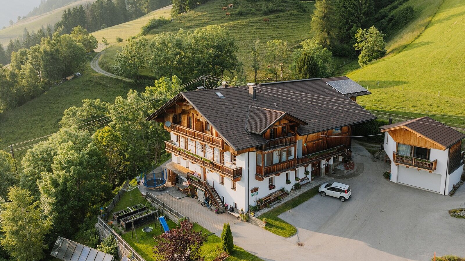 The farmhouse with balconies, a children's playground, and a sunbathing lawn, nestled in a green hilly landscape.