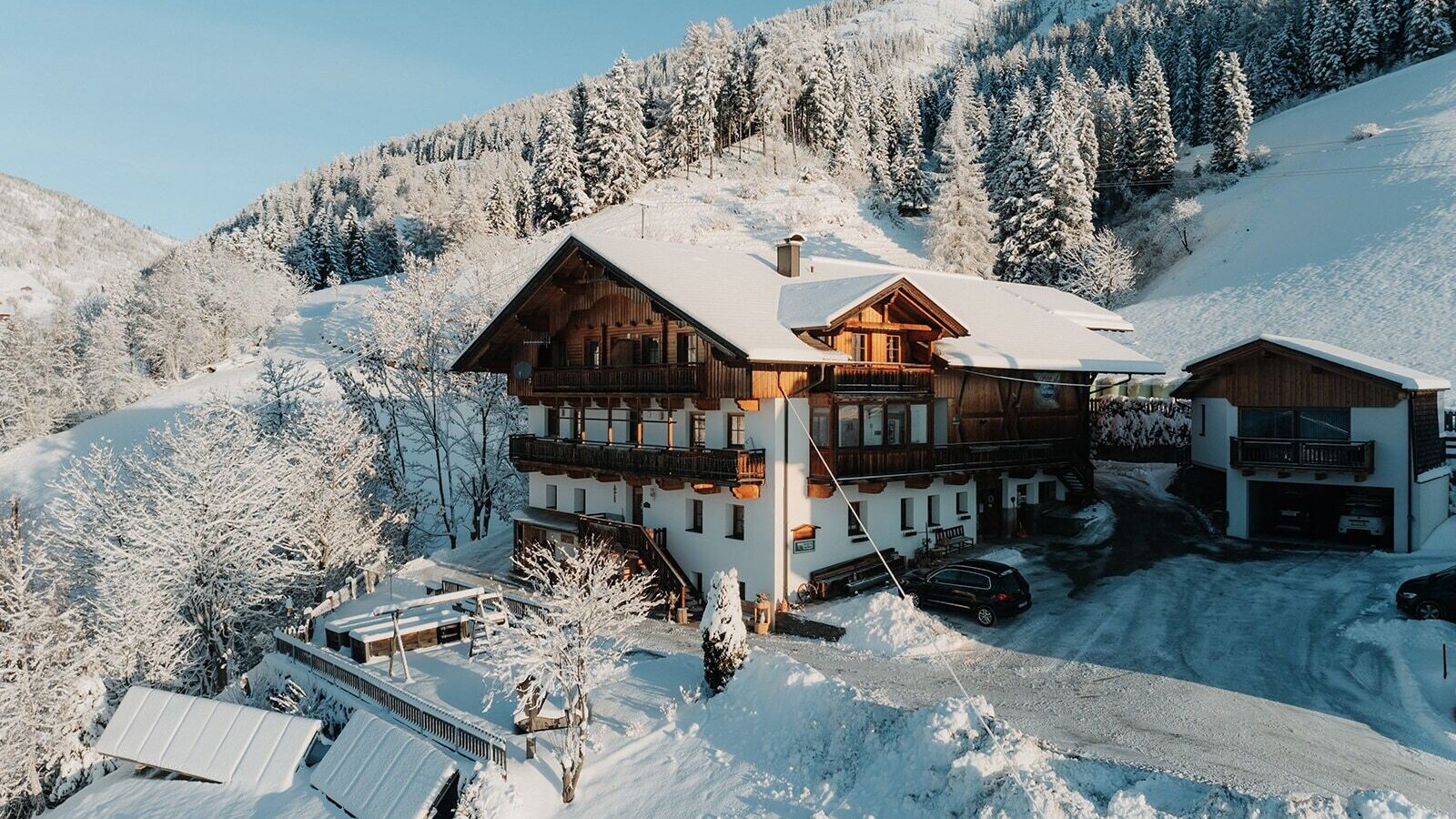 The farmhouse in winter with balconies and a children's playground on the terrace, surrounded by snowy mountains.