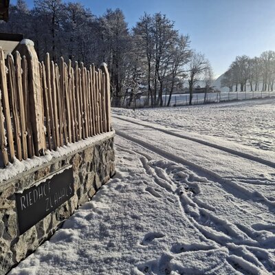 Entrance to the Riedhof-Zuahaus with stone wall, wooden fence, and snow-covered path on a sunny winter day.