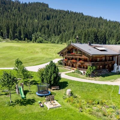 The idyllic farmhouse of the apartments, featuring a spacious green garden with a children's playground set against a dense forest backdrop.