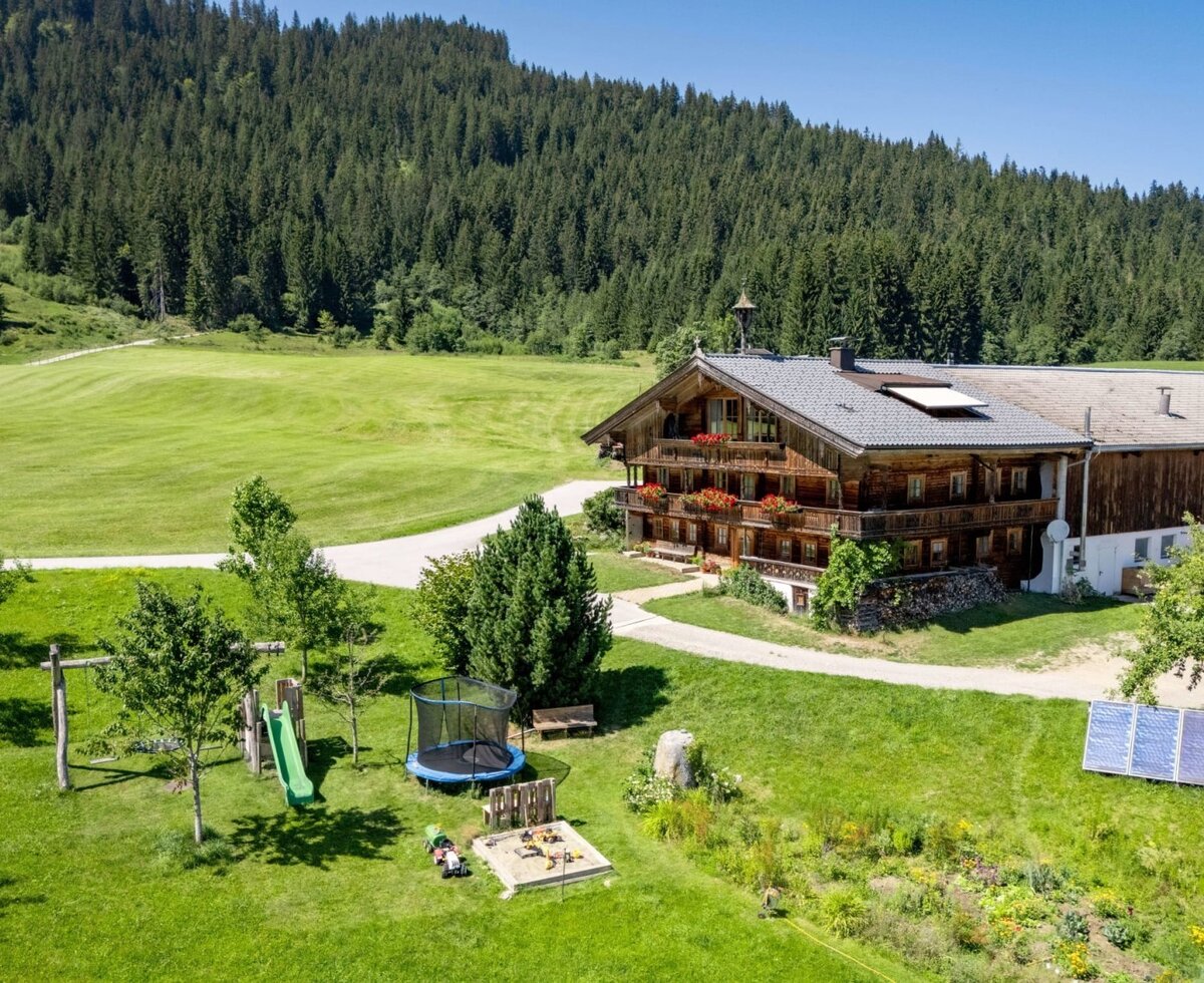 The idyllic farmhouse of the apartments, featuring a spacious green garden with a children's playground set against a dense forest backdrop.