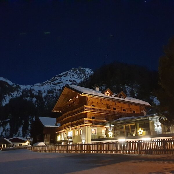 The Farm House exterior at night, featuring traditional wooden architecture, an illuminated facade, and a glass-enclosed area, set against a backdrop of snow and mountains.