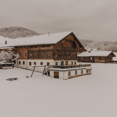 The snow-covered Ferienwohnung with a wooden facade, a terrace, and a playground, surrounded by a winter landscape.