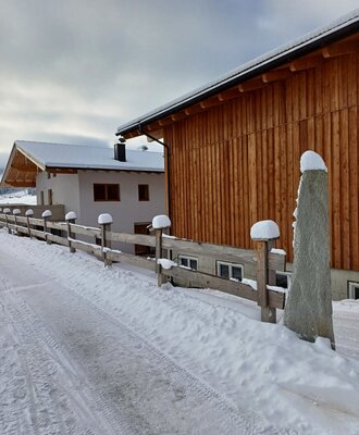 The farmhouse exterior in winter, featuring buildings with wooden and light-colored facades, a snow-covered path, and a wooden fence.