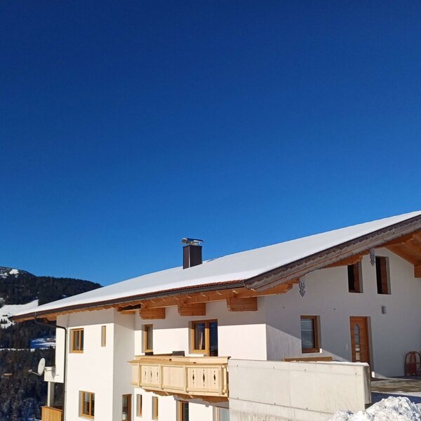 The Farm House exterior with its white facade, wooden balcony, and snow-covered gabled roof, surrounded by a snowy mountain landscape.