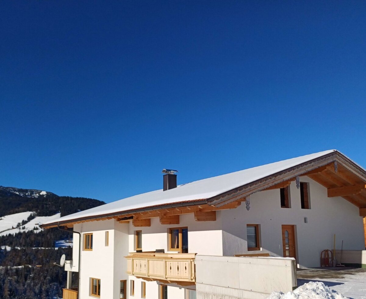 The Farm House exterior with its white facade, wooden balcony, and snow-covered gabled roof, surrounded by a snowy mountain landscape.
