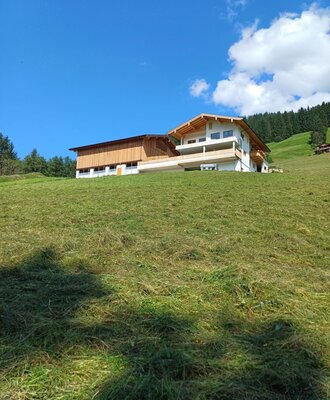 The farm house features a main building with balconies and an adjoining wooden barn, situated on a green hillside with surrounding forest.
