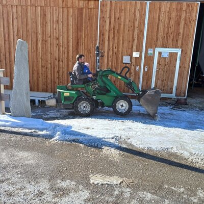 A green compact tractor with a man and child parked beside the wooden farm house, offering a glimpse into daily farm activities.