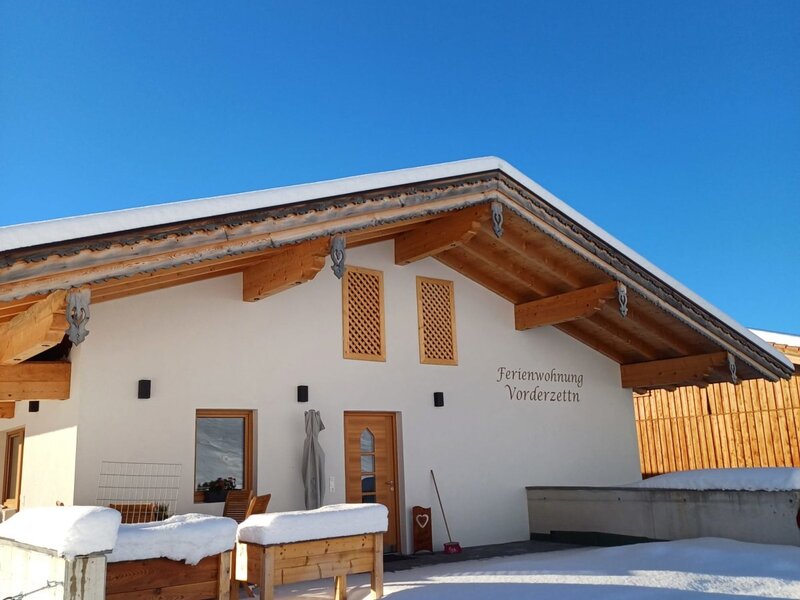 The winter exterior of the 'Ferienwohnung Vorderzettla' farmhouse, covered in snow under a clear blue sky.