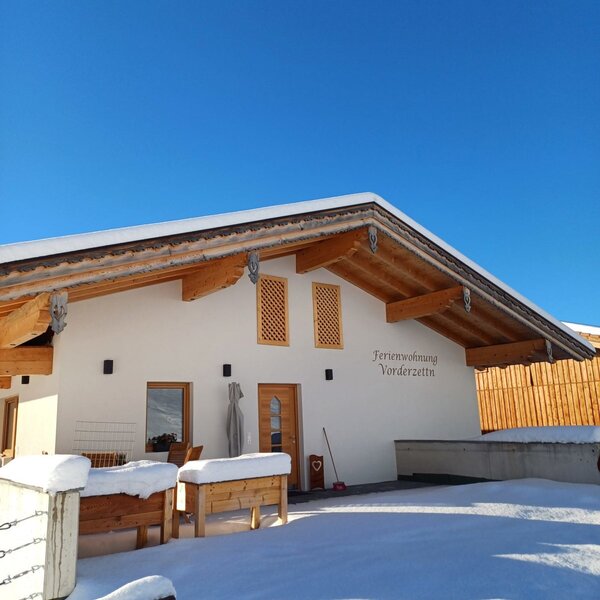 The winter exterior of the 'Ferienwohnung Vorderzettla' farmhouse, covered in snow under a clear blue sky.